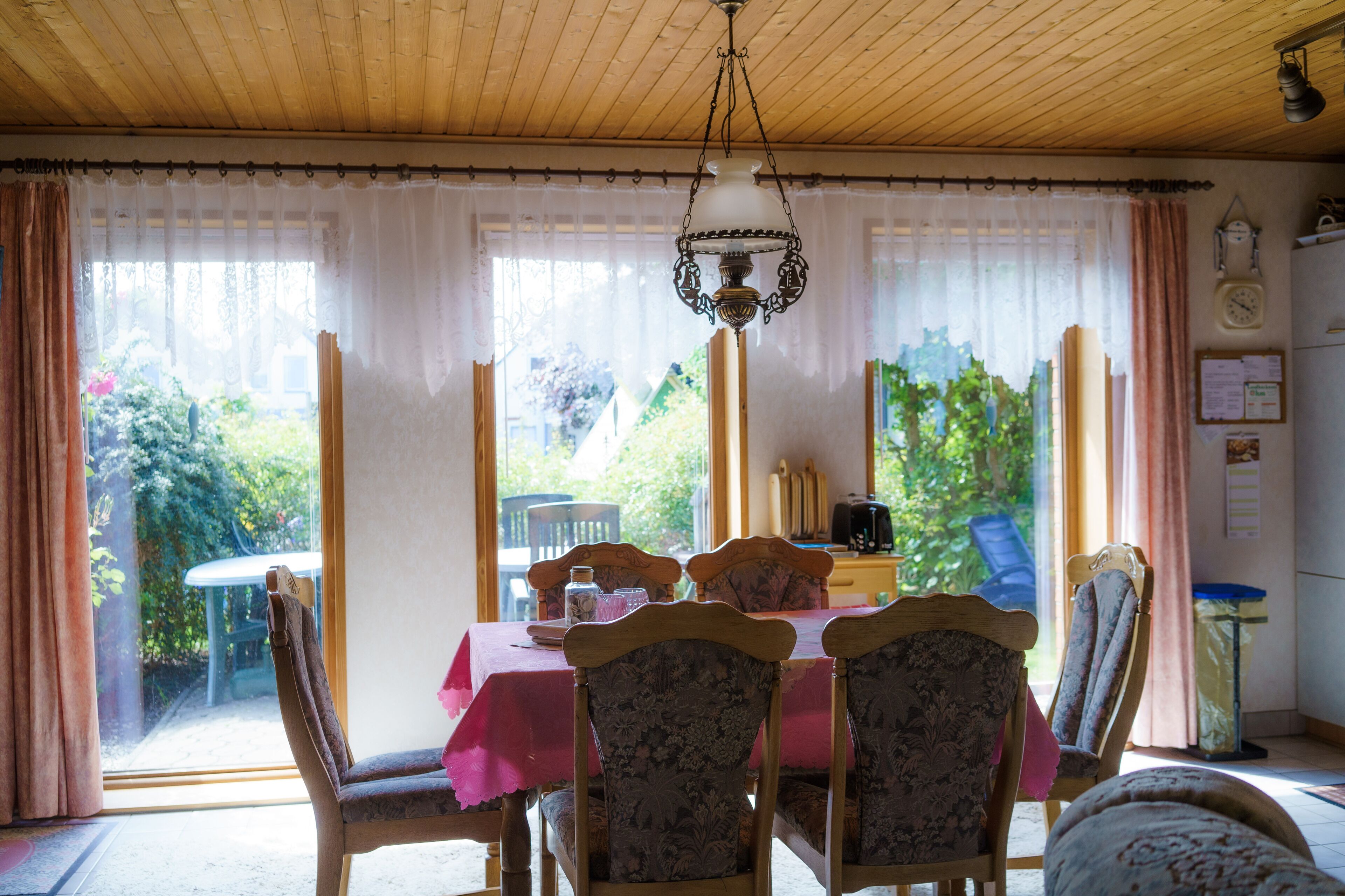A dining area with a table set for four, chairs, a chandelier, and a view of the garden through large windows.