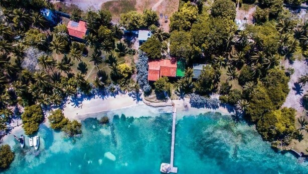 Vista aerea di una proprietà costiera con spiaggia, acque turchesi limpide e un molo che si protende verso il mare.