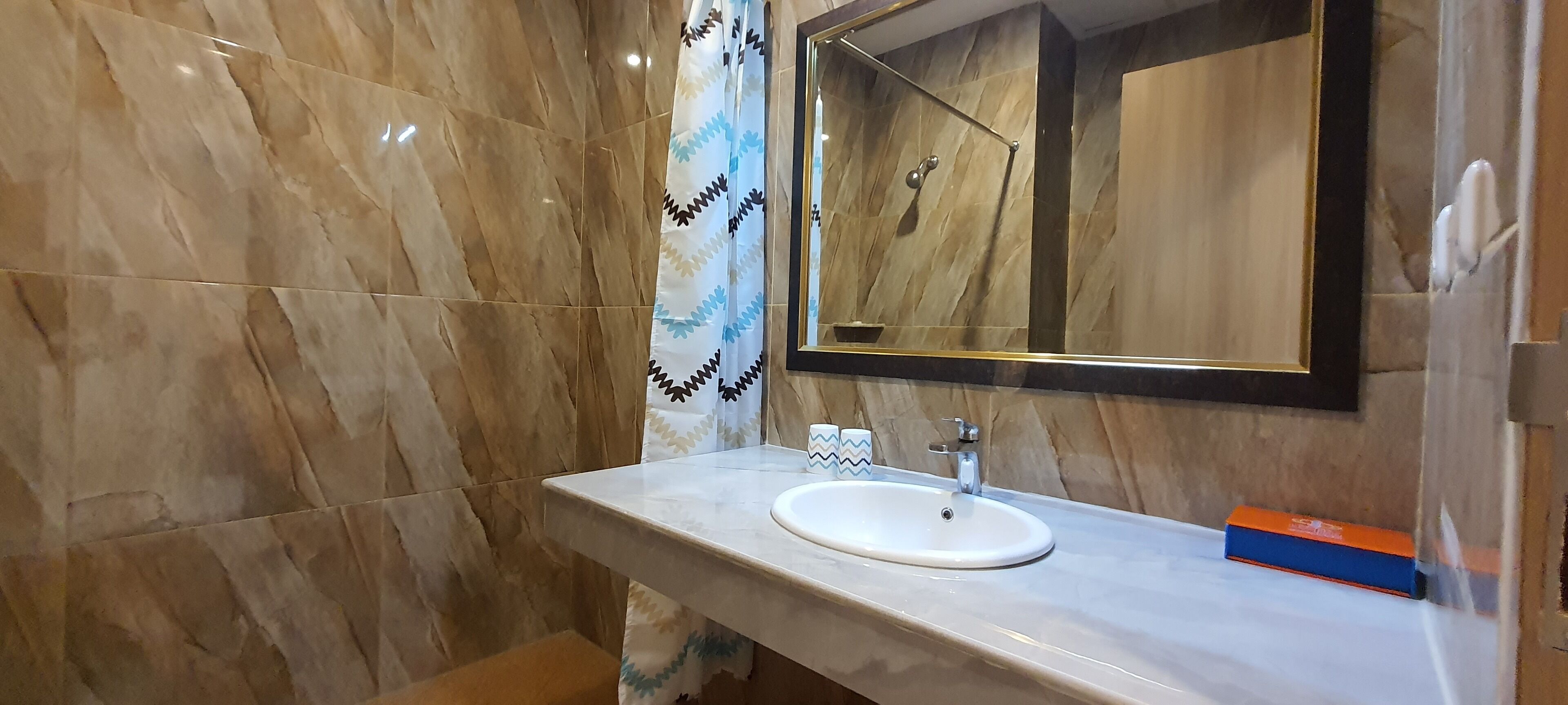 A bathroom with a marble floor, a white sink, a mirror with a gold frame, and a shower area.