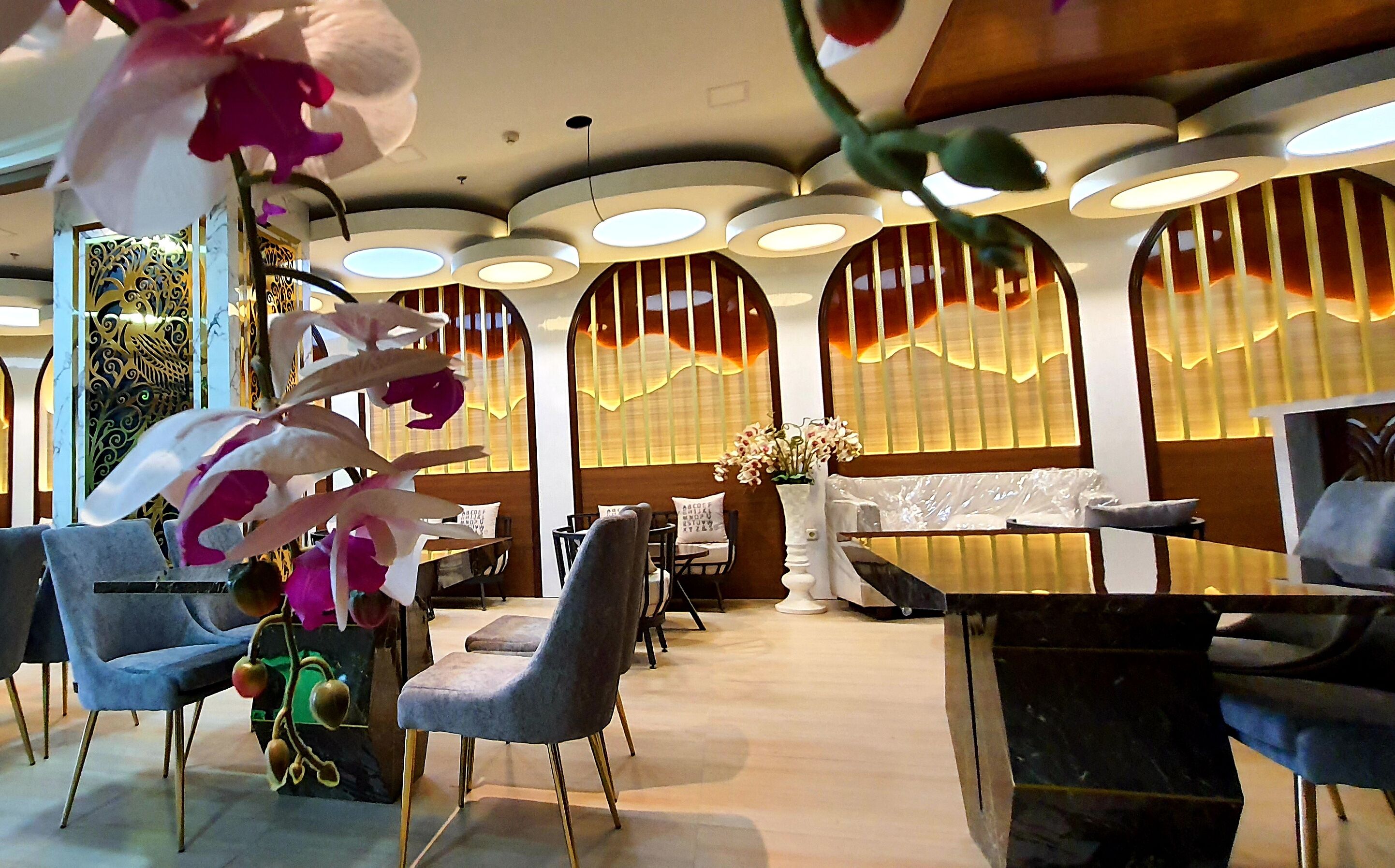 A restaurant interior with round tables, grey upholstered chairs, and decorative plants.