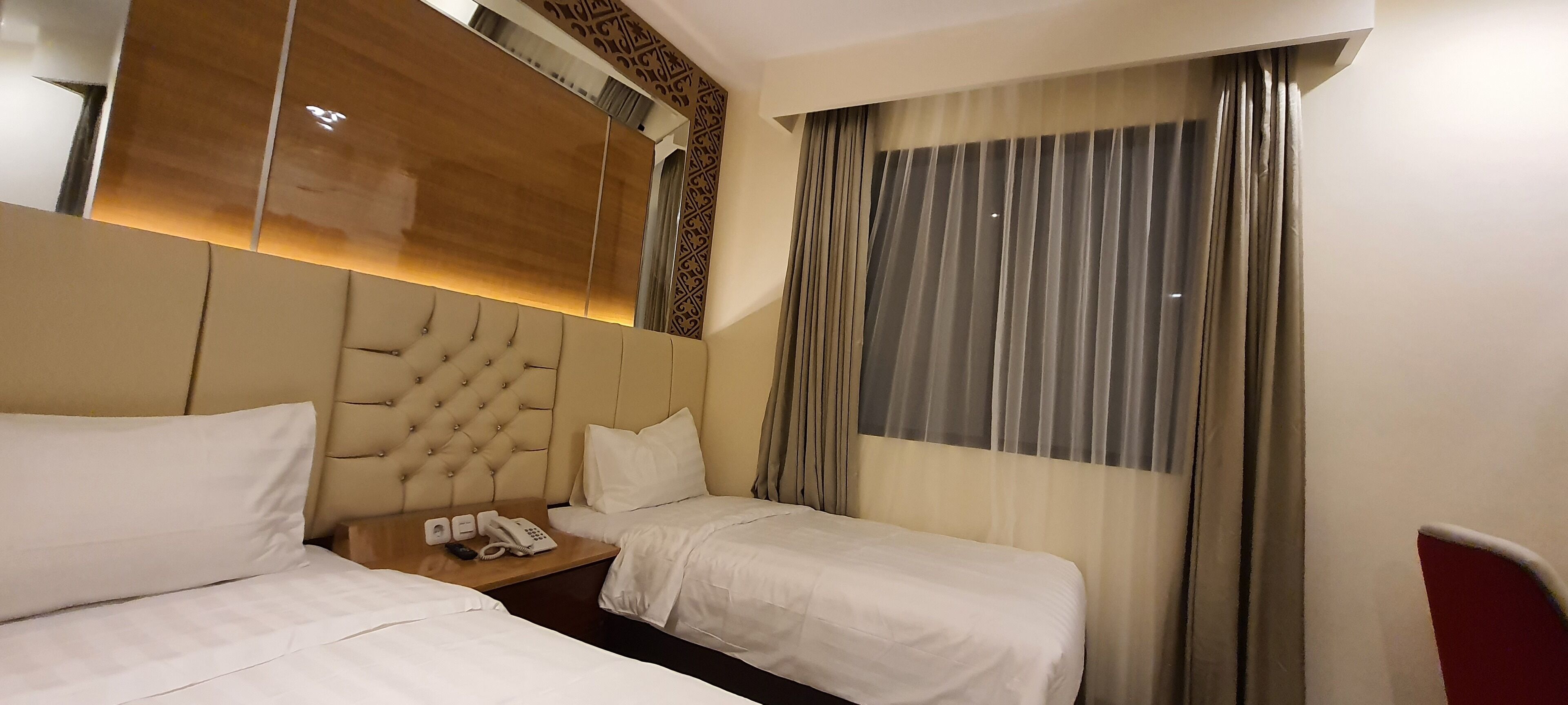 A hotel room with two beds, a wooden headboard, a bedside table with a phone and a lamp, and a large window with curtains.