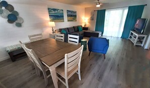 A living room with a wooden dining table, white chairs, a blue armchair, a sofa, and a TV stand.