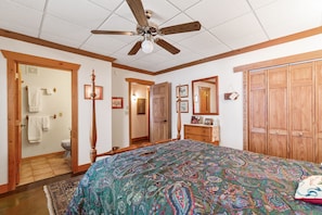 Une chambre à coucher avec un grand lit, une armoire en bois, un ventilateur de plafond et une salle de bain visible à travers une porte ouverte.
