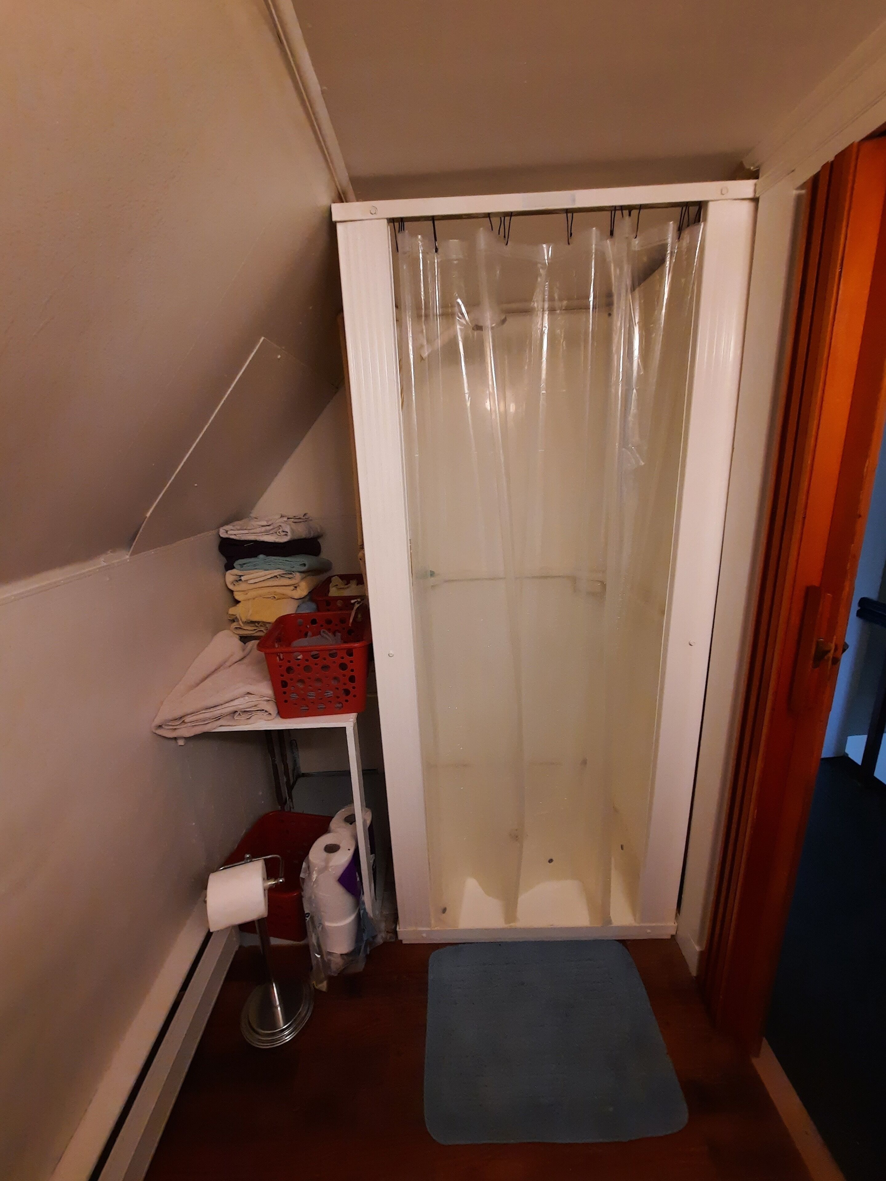 Une petite salle de bain étroite, avec un rideau de douche, une étagère où sont pliées des serviettes, un panier à linge rouge et un tapis bleu sur le sol.