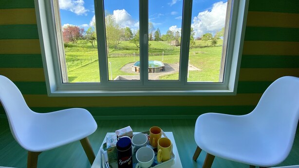 Een kamer met een groot raam dat uitkijkt op een groen landschap, met twee witte stoelen en een tafel waarop een dienblad staat met diverse spullen, waaronder een Senseo koffiezetapparaat, kopjes en een doos.
