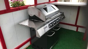 Een barbecue van roestvrij staal met vier branders en een bijbehorend bijzettafeltje, geplaatst tegen een witte muur met rode accenten.