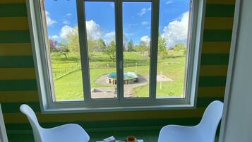 Ein Zimmer mit einem großen Fenster mit Blick auf eine Rasenfläche, auf der sich eine runde Struktur, zwei weiße Stühle und ein Tisch mit verschiedenen Gegenständen befinden.