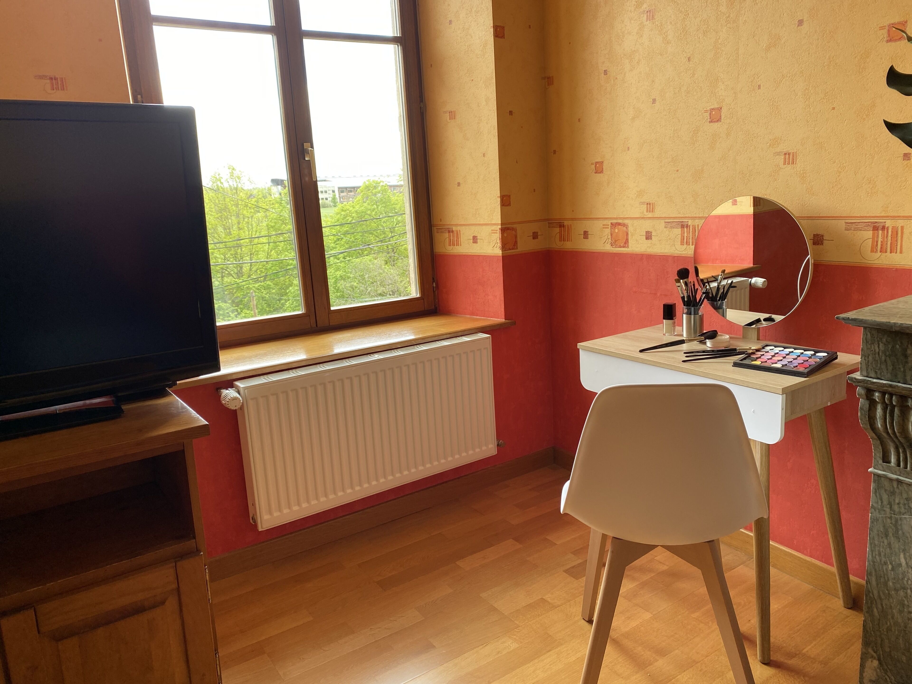 Una stanza con televisione su un supporto in legno, una scrivania bianca con specchio, cosmetici e una sedia.