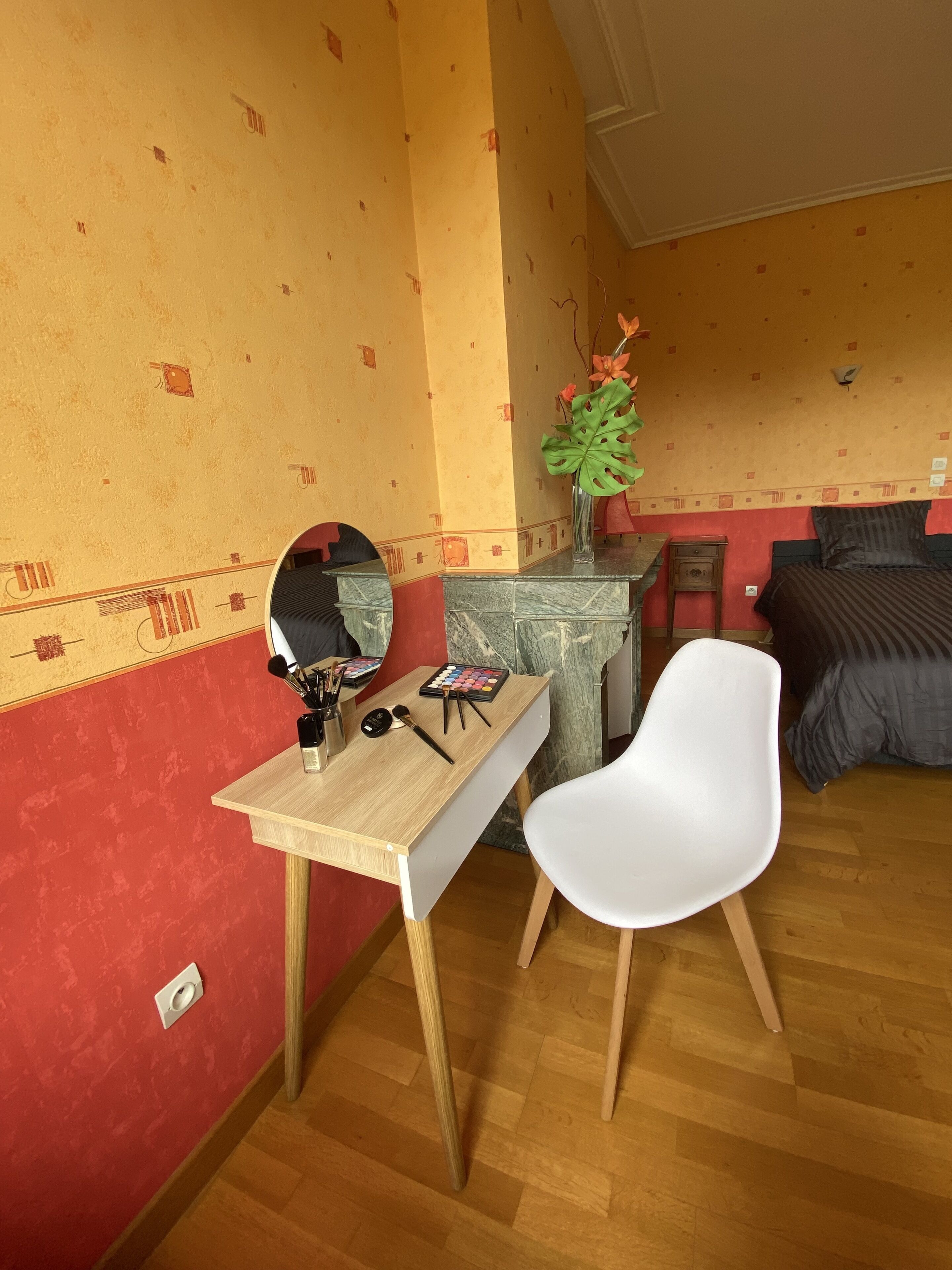 Ein Zimmer mit einem Holztisch, einem weißen Stuhl, einem Bett mit dunkler Bettwäsche, einem Spiegel und einer Vase mit Blumen.