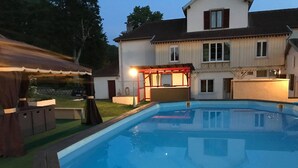 Een zwembad met een houten terras, een barbecueplek, en een huis met balkon op de achtergrond.