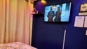 Une chambre d’hôtel avec un lit, un téléviseur fixé au mur, un rideau et une petite étagère avec quelques objets.