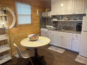 A compact kitchen with white cabinets, a microwave, a stove, and a dining area with a table and chairs. - Pet Friendly Separate Fee, enjoy fenced in yard, outdoor shower, fire pit (Holden beach)