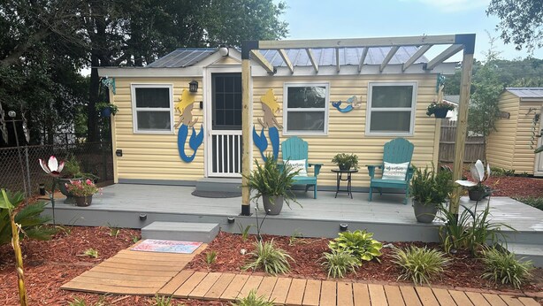 A small, single-story building with a deck, potted plants, and outdoor seating. - Pet Friendly Separate Fee, enjoy fenced in yard, outdoor shower, fire pit (Holden beach)