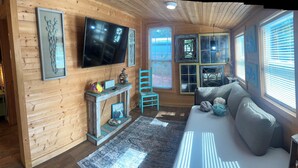 A cozy cabin interior with a wooden wall, a mounted TV, a sofa with cushions, a small table with a lamp, and a window with blinds. - Pet Friendly Separate Fee, enjoy fenced in yard, outdoor shower, fire pit (Holden beach)