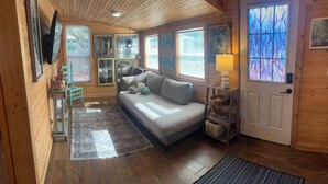 A cozy cabin interior with a sofa, wooden walls, a television, and a window with a stained glass view. - Pet Friendly Separate Fee, enjoy fenced in yard, outdoor shower, fire pit (Holden beach)
