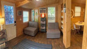 A cozy cabin interior with two grey sofas, a dining table, a lamp, and artwork on the walls. - Pet Friendly Separate Fee, enjoy fenced in yard, outdoor shower, fire pit (Holden beach)