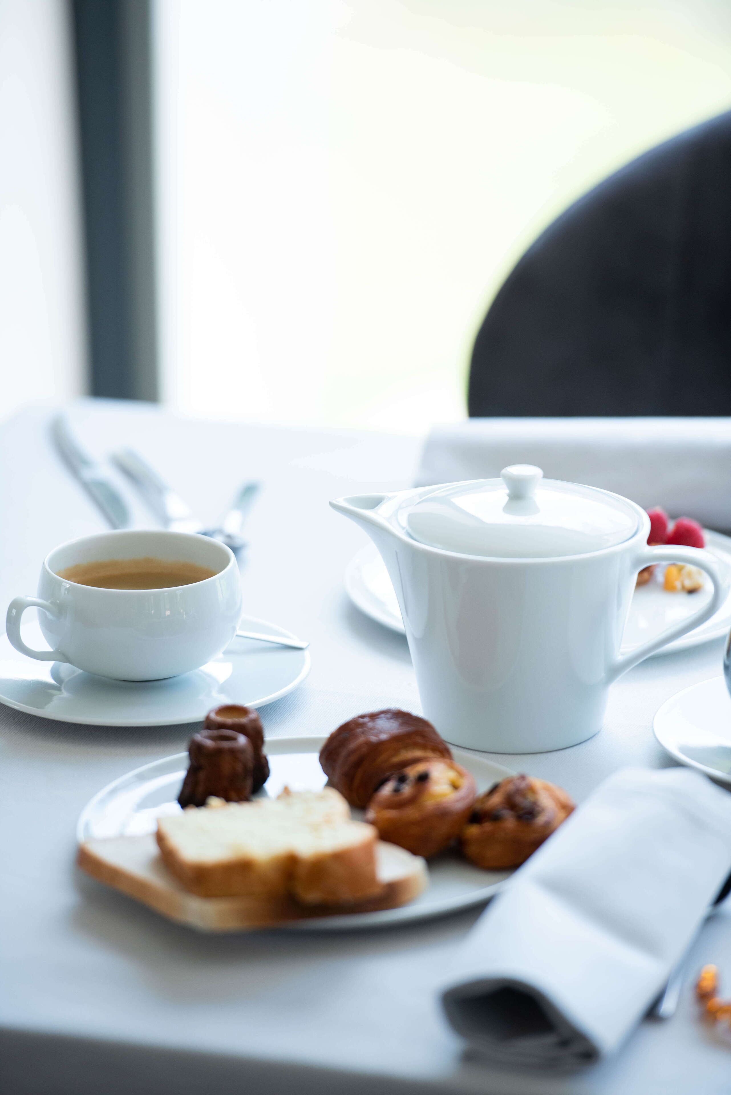 A table with a white cup of coffee, a white teapot, and pastries on a plate.