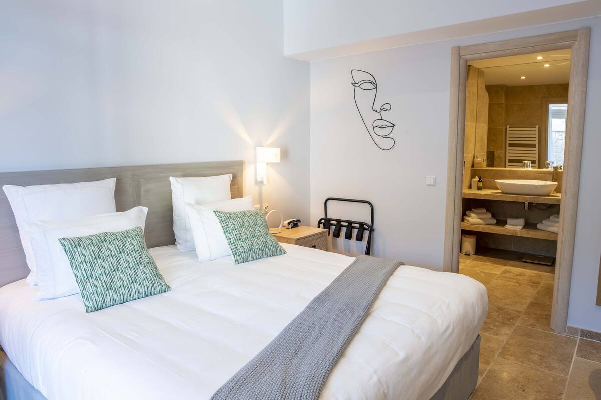 Une chambre d’hôtel avec un grand lit, une table de chevet, une lampe, un porte-serviettes et une salle de bain visible à travers une porte ouverte.