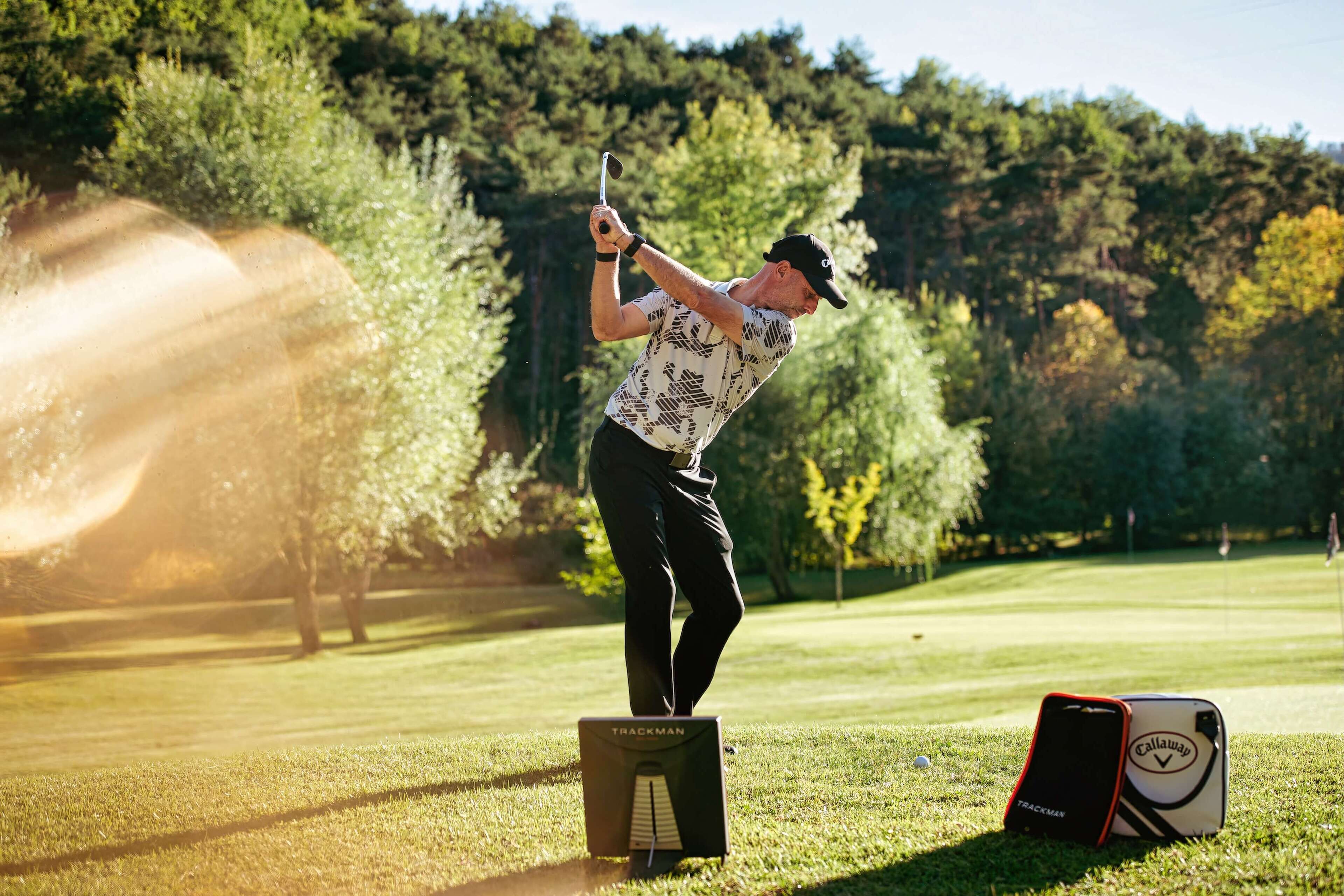 A golfer in mid-swing on a golf course with trees in the background.