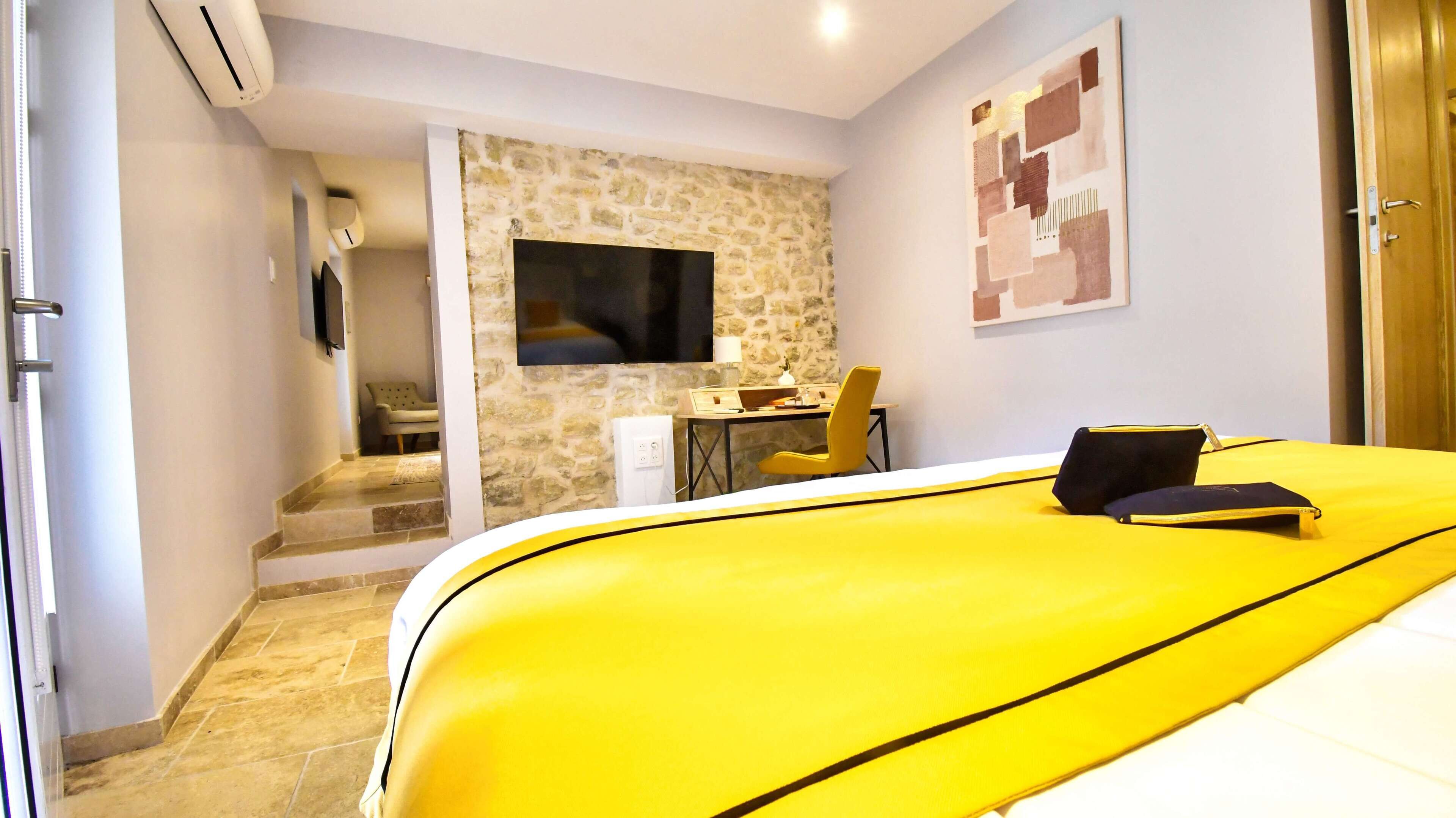 A hotel room with a yellow bed, a stone wall, a flat-screen TV, a desk with a chair, and a staircase leading to another room.