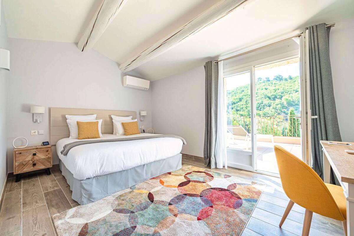 Une chambre à coucher comprenant un lit, une table de chevet, un bureau et donnant sur un espace verdoyant.