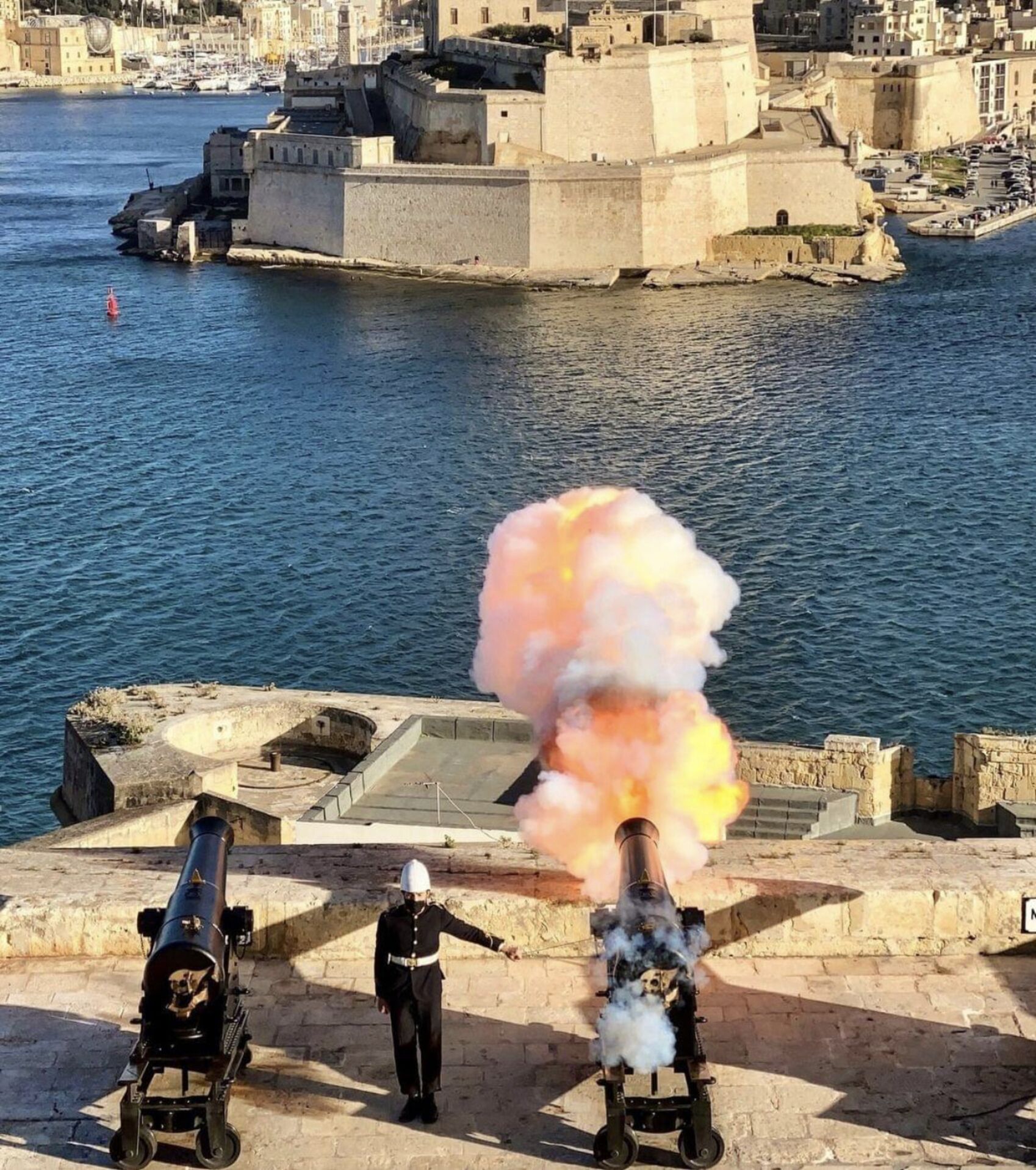 A historical coastal fortification with cannons, a clear view of the sea, and a cityscape in the background.