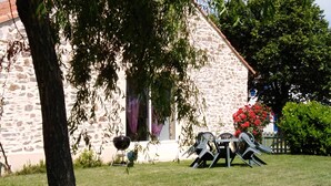 A stone building with a stone wall, a window with purple curtains, and a table with chairs in the yard.