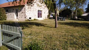 A stone house with a red-tiled roof, a small front porch, and a fenced yard with a tree and a table with chairs.