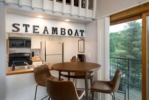 Une cuisine moderne avec une enseigne « STEAMBOAT », une table ronde en bois entourée de quatre chaises, et une vue sur la verdure depuis un balcon.