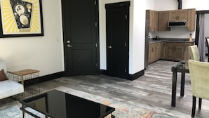 Una sala moderna con una mesa de centro de vidrio negro, un sofá y un tapete colorido. Junto a ella, una cocina con gabinetes de madera y una estufa negra.