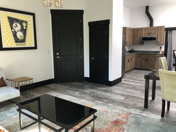 Una sala moderna con una mesa de centro de vidrio negro, un sofá y un tapete colorido. Junto a ella, una cocina con gabinetes de madera y una estufa negra.