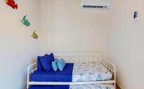 A single-bed room with a white metal frame, a blue mattress, and matching blue pillows. There is a wall-mounted air conditioning unit above the bed.