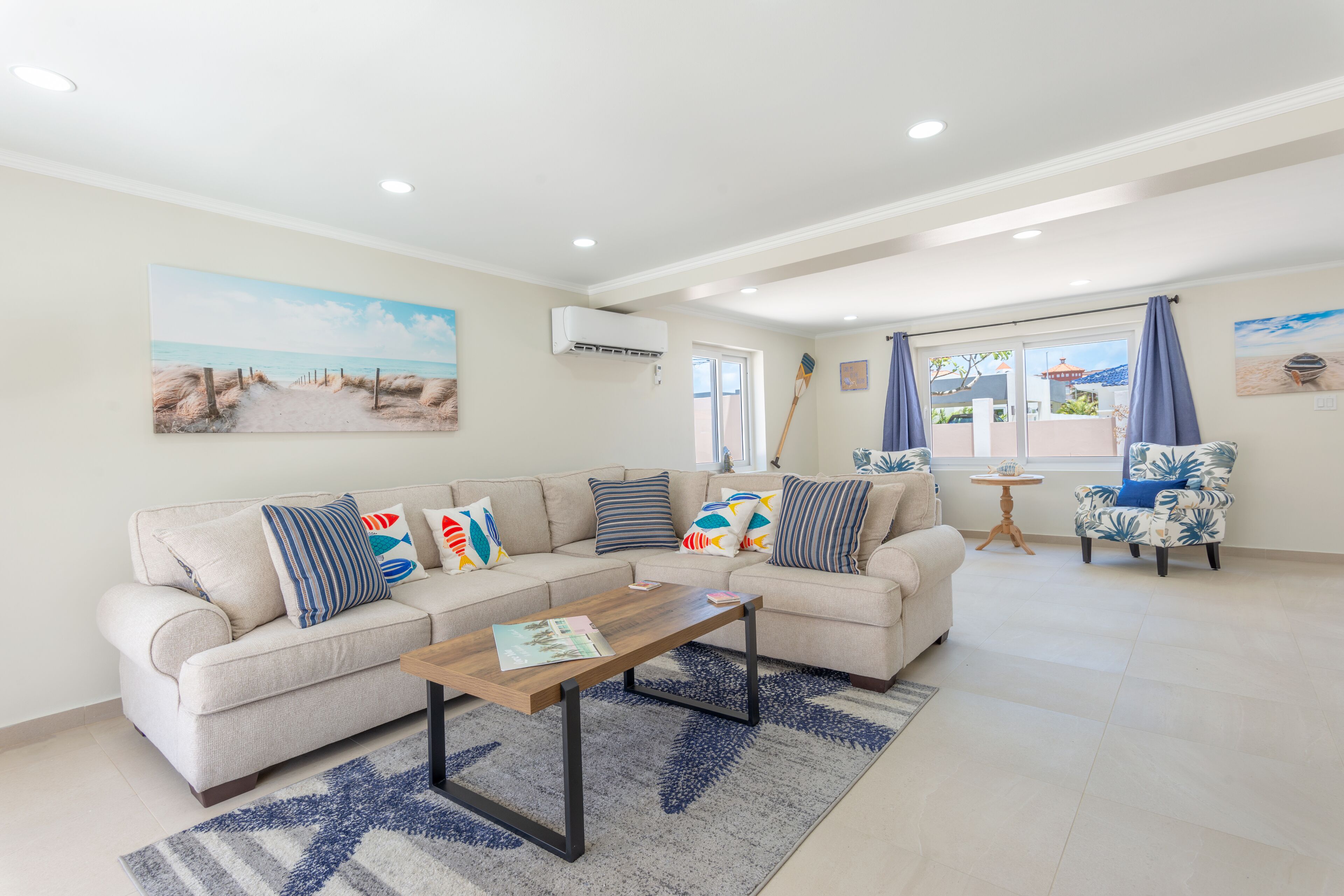 Un salón con un sofá color beige, una mesa de centro de madera y un tapete estampado. Hay dos sillones con cojines azules, una mesita redonda y un cuadro con una escena de playa en la pared.