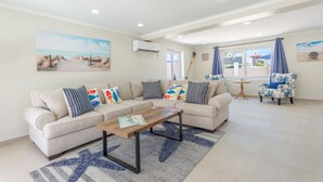 Un salon avec un canapé beige, une table basse en bois et un tapis à motifs. On y trouve deux fauteuils avec des coussins bleus, une petite table ronde et un tableau représentant une scène de plage accroché au mur.