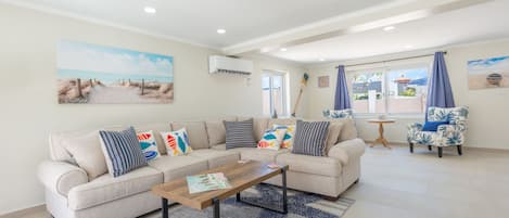 A living room with a beige sofa, a wooden coffee table, and a patterned rug. There are two armchairs with blue cushions, a small round table, and a painting of a beach scene on the wall.