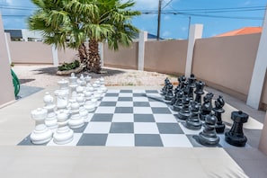 Un grand jeu d’échecs installé en plein air, sur une terrasse, avec un palmier et un mur blanc en arrière-plan.