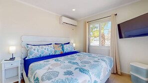 A bedroom with a bed, nightstand, lamp, air conditioning unit, window with curtains, and a television.