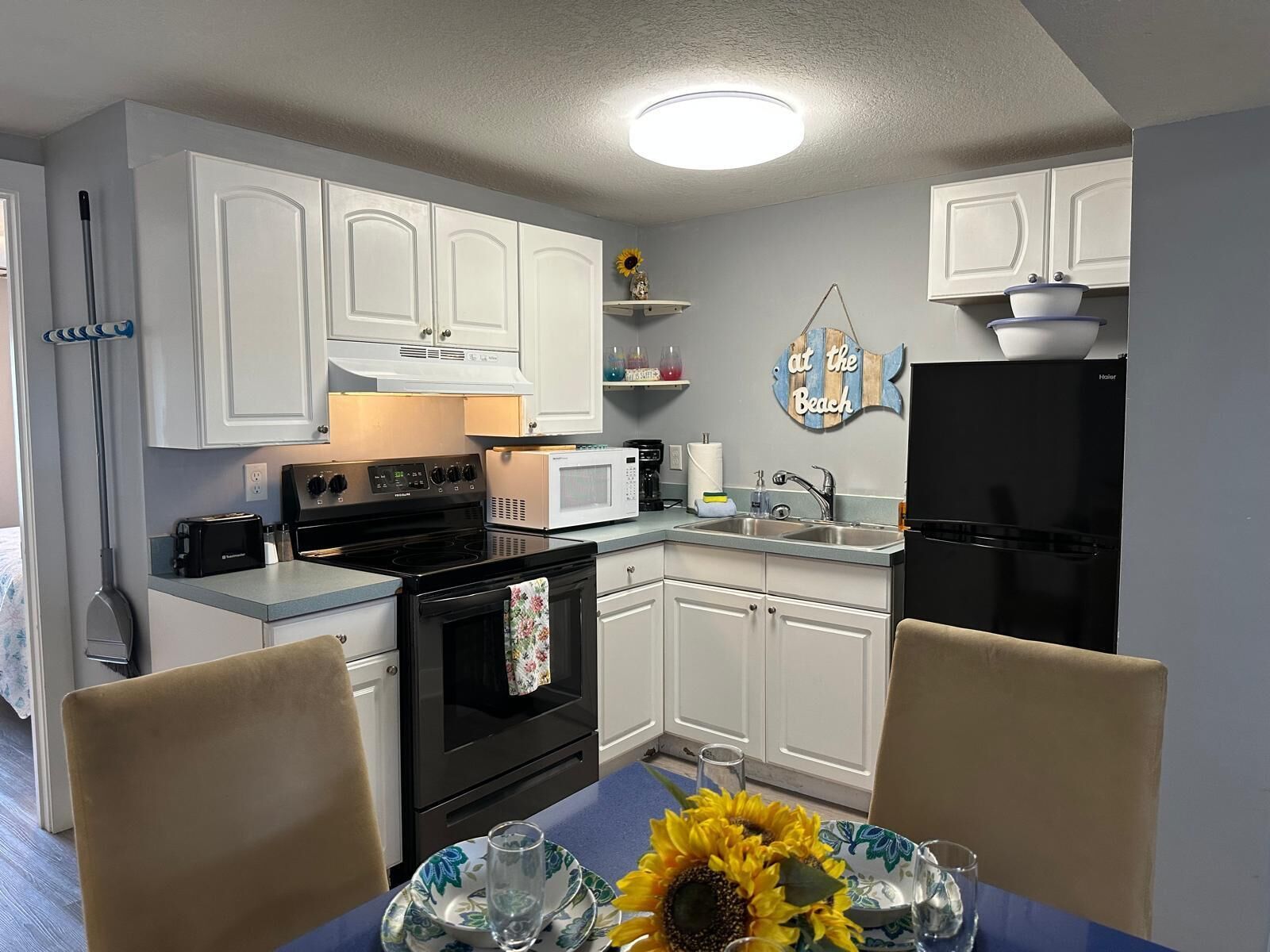 A compact kitchen with white cabinets, a black stove, a microwave, and a refrigerator. There is a dining table with a blue tablecloth and a sunflower centerpiece.