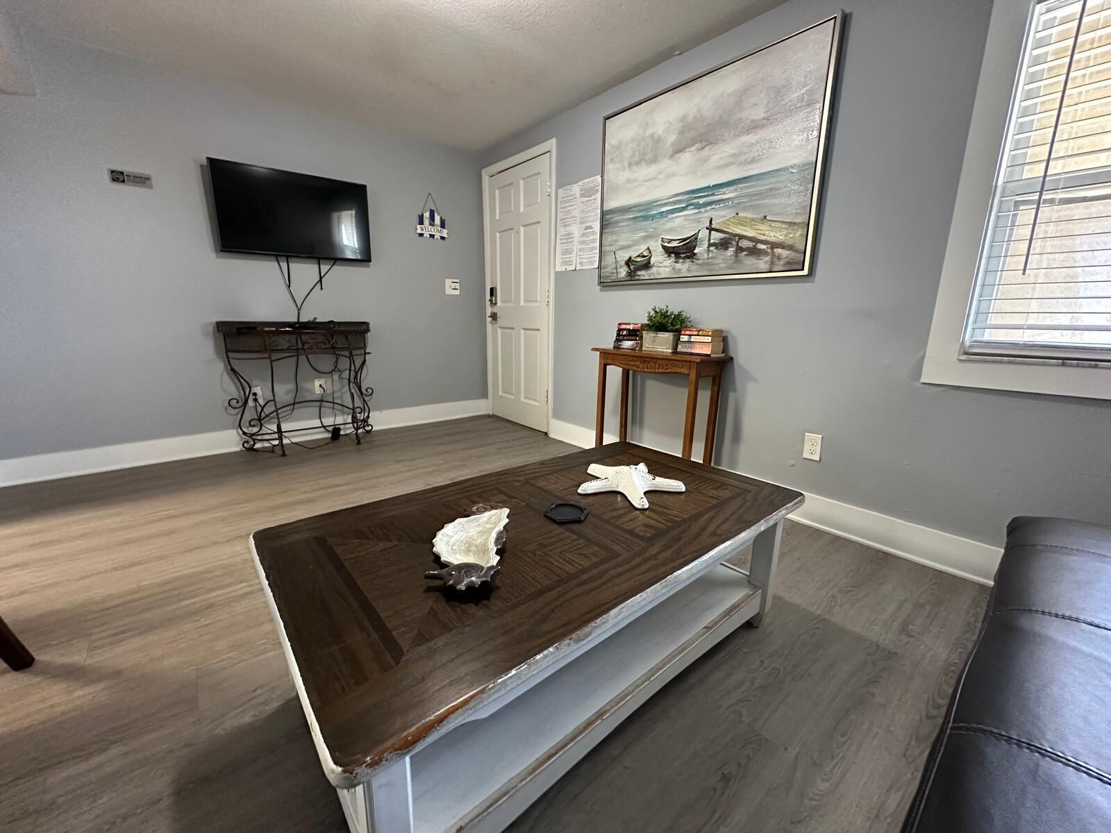 A living room with a dark wooden coffee table, a flat-screen TV, a painting of boats, and a small wooden side table with books and a plant.