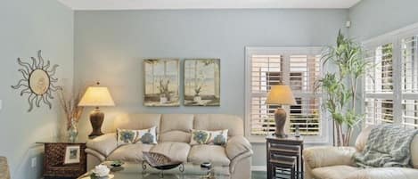 A living room with a beige sofa, a glass coffee table, and two lamps. There are wall art pieces and a window with blinds.