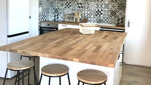 Une cuisine dotée d’un grand îlot central en bois, de tabourets en métal noir, d’un luminaire suspendu, d’un pare-feu décoratif et d’armoires en bois.