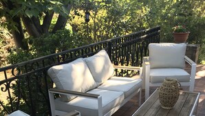 Un espace patio aménagé avec des meubles d’extérieur blancs, comprenant un canapé et deux fauteuils, une table basse en bois et une plante en pot.