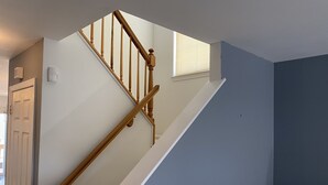 Uma escadaria com corrimãos de madeira e rodapé branco, que leva a um quarto com porta e janela com persianas.