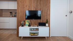 Ein modernes Wohnzimmer mit einem Flachbildfernseher an einer Holzwand, einem weißen Konsolentisch mit Büchern und Dekoration sowie einer Küche im Hintergrund.