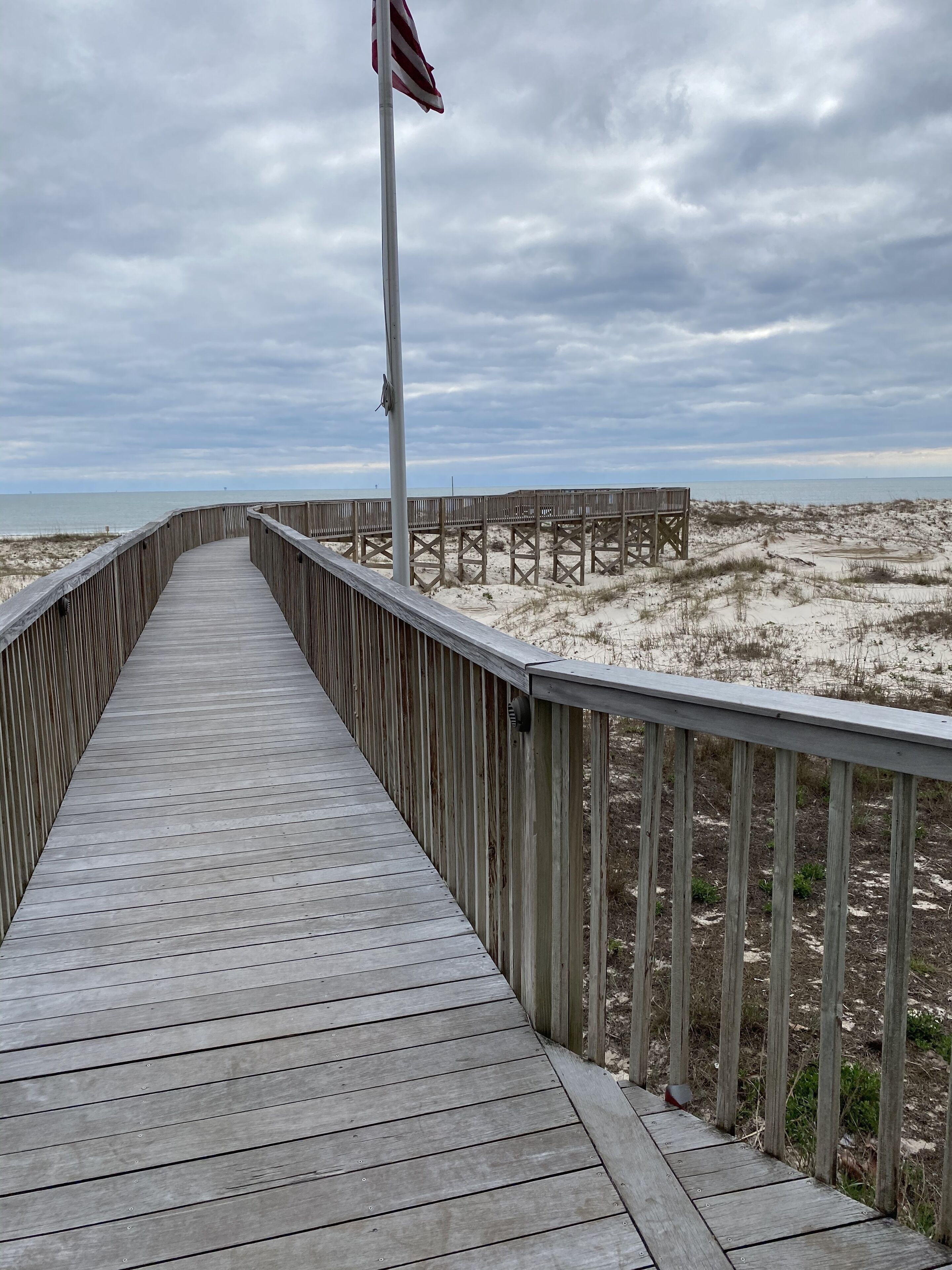 A wooden boardwalk leading to a sandy beach area with a flagpole in the distance.