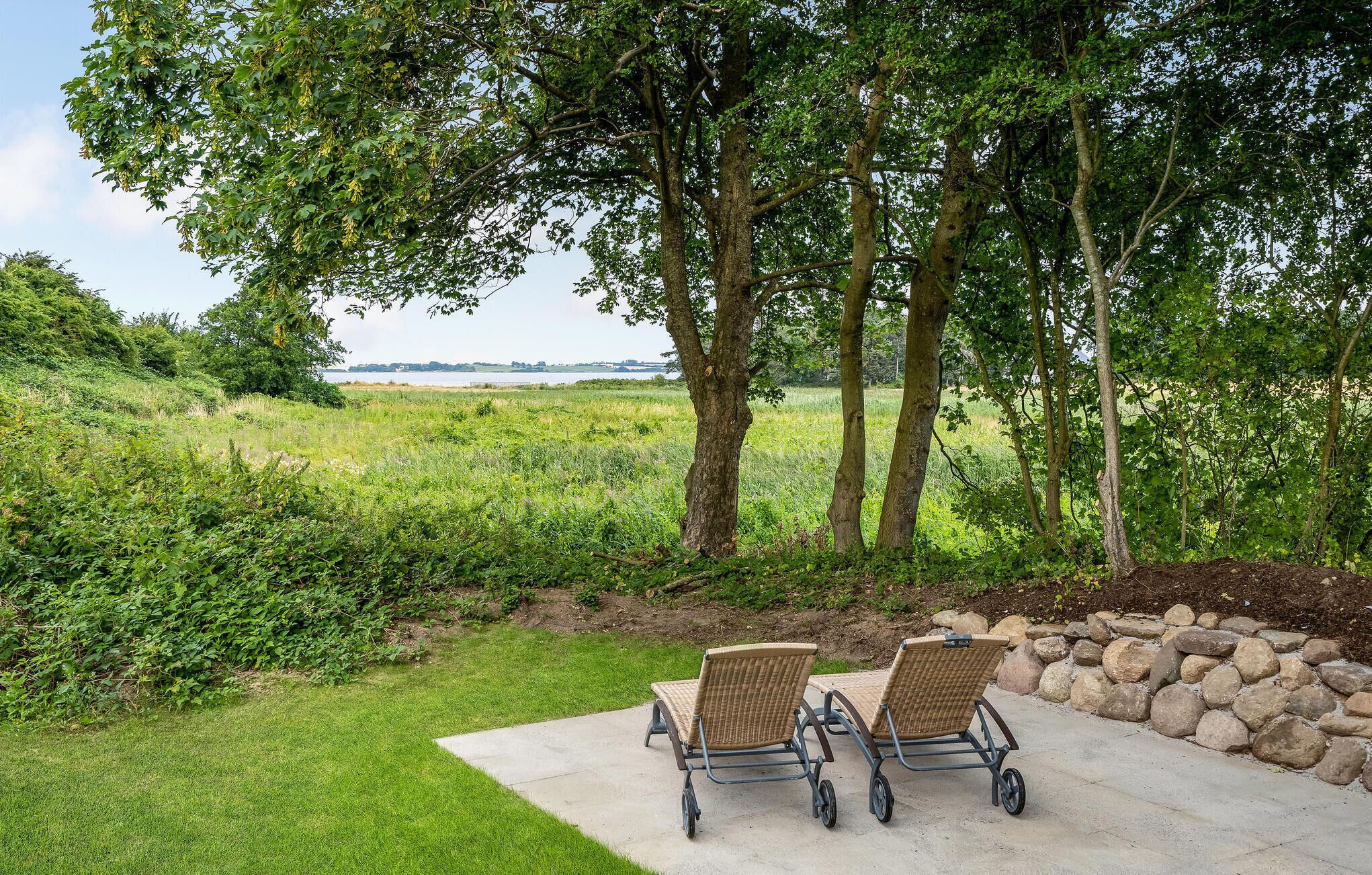 Zwei Stühle im Freien auf einer Beton-Terrasse mit Blick auf ein grasbewachsenes Feld und Bäume.