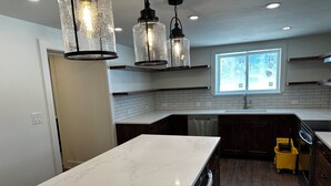 Una cocina moderna con encimeras de mármol blanco, armarios de madera oscura y un lavavajillas empotrado. Hay luces colgantes de diseño rústico, un ventanal que proporciona luz natural y un carrito de limpieza amarillo.