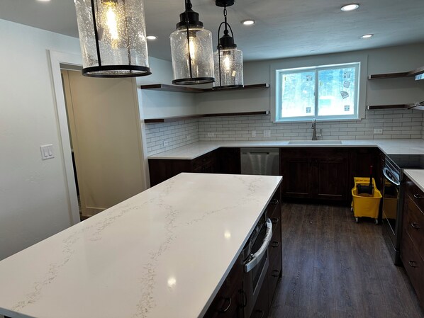 A modern kitchen with white marble countertops, dark wooden cabinets, and a built-in dishwasher. There are pendant lights with a rustic design, a window providing natural light, and a yellow cleaning cart.