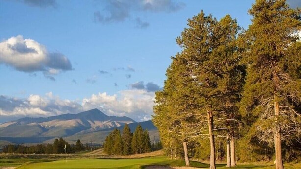 Un campo de golf con una bandera, árboles y montañas al fondo.
