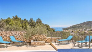 Area piscina con pareti in pietra e poltrone da relax blu, affacciata su una cittadina costiera e il mare.